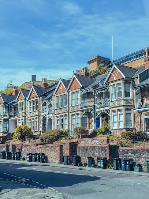 A row of Victorian-style terraced houses with ornate white ironwork balconies and large bay windows, situated behind a brick wall with small gardens and bushes in front. The properties are located on 