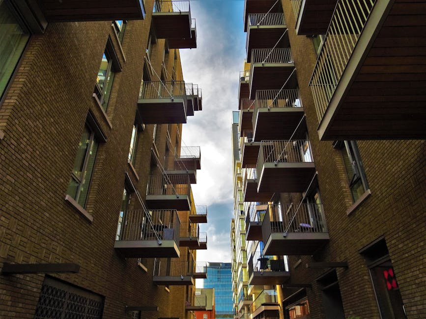 View looking up between two brown brick apartment buildings with multiple small balconies featuring metal railings. Some balconies have visible items such as outdoor furniture and plants. The sky is partly cloudy with patches of blue visible through the narrow space between the buildings. The perspective emphasizes the height and close proximity of the structures, suggesting an urban setting suitable for house removals. This scene relates to furniture transport and loading during a home relocation process, as carried out by Movers Hammersmith, who handle removals in tight city spaces and apartment blocks.