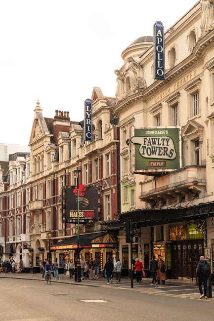The exterior of the Lyric Theatre in Hammersmith features a historic building with ornate architectural details and multiple vertical signage banners displaying the theatre's name. Adjacent to the theatre is the Apollo cinema with a prominent blue vertical sign. The street scene includes pedestrians walking along the pavement, some of whom are nearby a shopfront with illuminated signage. The environment suggests a busy urban area suitable for house removals and furniture transport, with potential for loading or unloading furniture and boxes. The lighting is natural, possibly during daytime, highlighting the building facades and street activity, making it relevant for home relocation services offered by Movers Hammersmith focusing on narrow street access and local moving logistics.