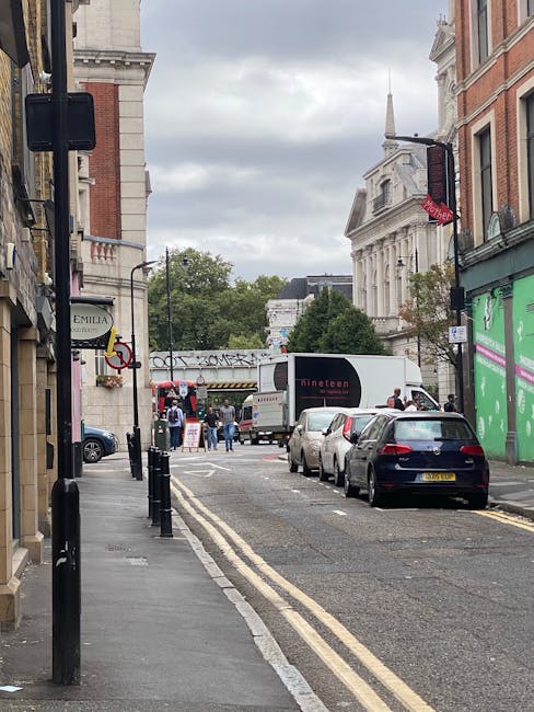 A narrow street scene in the Lyric Theatre area with a white delivery van parked near the curb, partially obstructing the view of a loading area behind it. Several cars are parked along the side of the street, with black bollards lining the pavement to prevent parking on the sidewalk. Pedestrians are walking on the pavement, some near a building façade with a green shopfront. Further down the street, a group of people are gathered near an entrance with a temporary structure or tent set up, indicating some activity related to home relocation or furniture transport. In the background, tall historic buildings with brick and stone facades are visible, along with a church steeple rising above the rooftops. The overcast sky provides diffused lighting, highlighting the scene's urban environment suitable for local house removals and moving services, as handled by Movers Hammersmith.