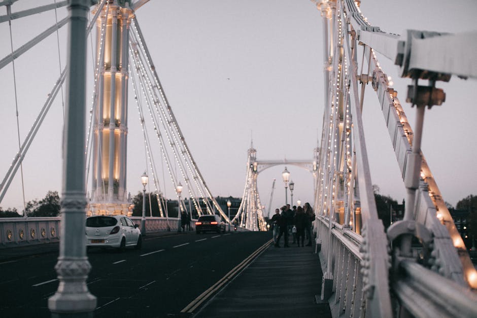 The image shows a view of a lit-up suspension bridge during dusk, with decorative string lights along the white metal framework of the bridge. Several vehicles, including a white car and a dark-colored car, are seen driving on the road, while a group of people are walking on the pavement, possibly involved in a home relocation or furniture transport process. The bridge features ornate lamp posts along its length, illuminating the area, and the background reveals a cloudy sky with trees faintly visible beyond the bridge. The scene captures the environment where movers from Movers Hammersmith may be executing loading or unloading activities, transporting furniture or packed boxes across or near the bridge for house removals in the Hammersmith area. Equipment such as trolleys, straps, or blankets is not visible in this specific image, but the overall setting reflects the logistics involved in packing and moving services associated with property relocation near Hammersmith Bridge, highlighting aspects of transportation and access during the process.