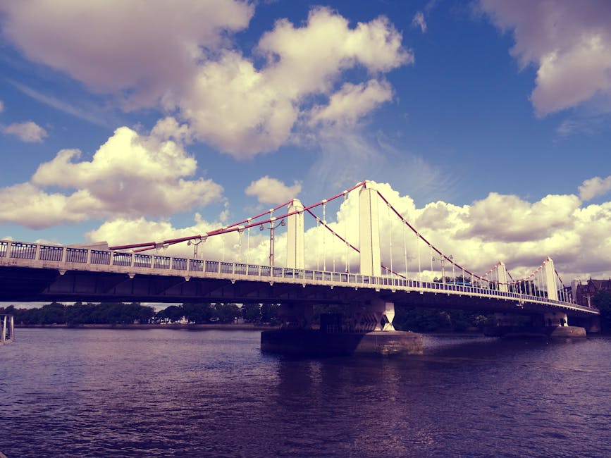 An exterior view of a large suspension bridge with white towers, red suspension cables, and a pedestrian walkway, spanning over a river with calm water. The sky above features scattered white clouds against a bright blue background. In the foreground, the riverbank is visible, with the base of the bridge supported by stone pillars. The area appears to be a staging point for home relocation or furniture transport, with no visible moving equipment or personnel in the image. The scene emphasizes the structural and scenic aspects of the bridge, relevant to the logistics of house removals near Hammersmith Bridge, as handled by Movers Hammersmith during a typical loading or unloading process.