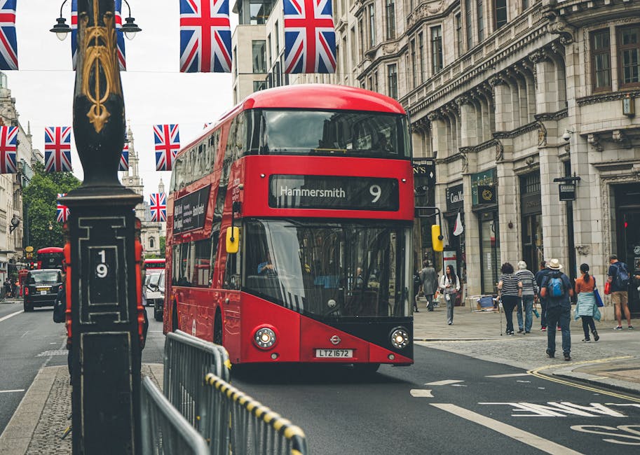 A red double-decker bus displaying the route number 9 and destination 'Hammersmith' is driving along a busy street in central London during daytime. The bus is approaching pedestrians crossing the road and is framed by Union Jack flags hanging above the street, which are attached to black lampposts. On the right side, a group of people are walking on the sidewalk near historic stone buildings with ornate architectural details. In the foreground, a metal barrier and a black signpost are visible near the bus lane, while the pavement is made of patterned stones. The scene captures typical city traffic with other vehicles visible further down the street. As part of home relocation or furniture transport logistics, professional movers such as Movers Hammersmith may use such routes for efficient house removals in the Hammersmith area, ensuring safe and timely transportation of household items.