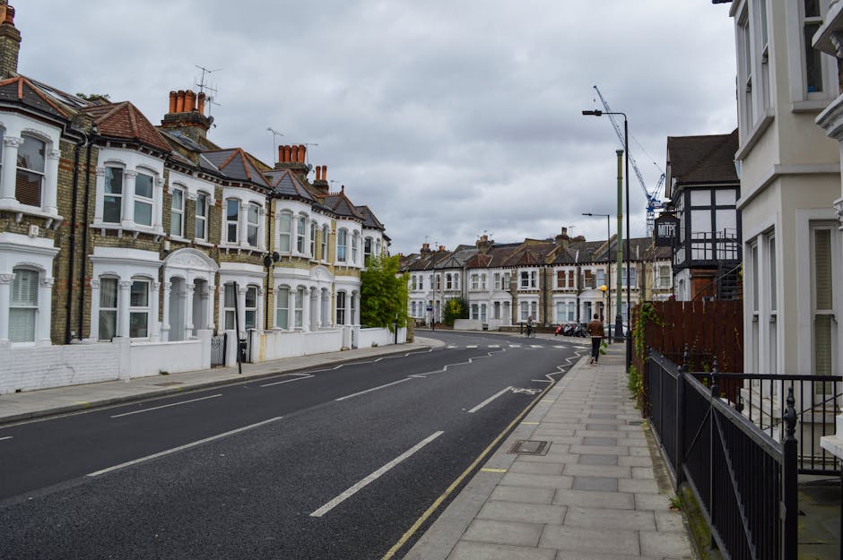 Photograph of a residential street in Hammersmith, showing traditional Victorian-style terraced houses with white facades, bay windows, and red-tiled roofs. The street features a paved sidewalk on the right side, bordered by a black metal fence and house entrances. A few pedestrians are walking along the pavement, and one person is visible near the middle distance. The road is empty of vehicles, with clear lane markings, and an overcast sky with gray clouds overhead. In the background, there are construction cranes indicating ongoing development nearby. This setting illustrates an urban environment suitable for house removals and furniture transport which nearby removals services like Movers Hammersmith handle, particularly during home relocation and packing and moving processes involving street access and vehicle loading or unloading.